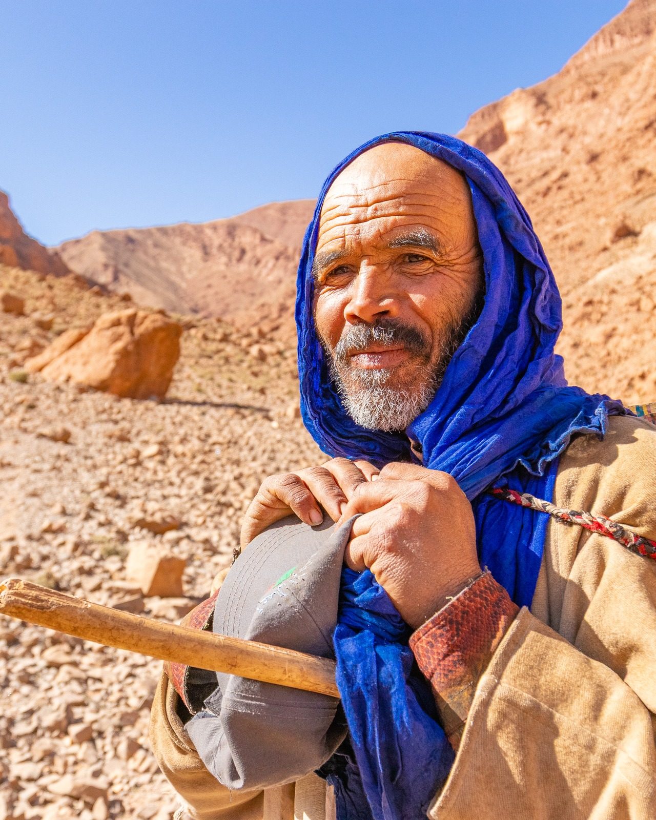 Portrait of an older man in a blue headscarf holding a cap, rocky hillside behind